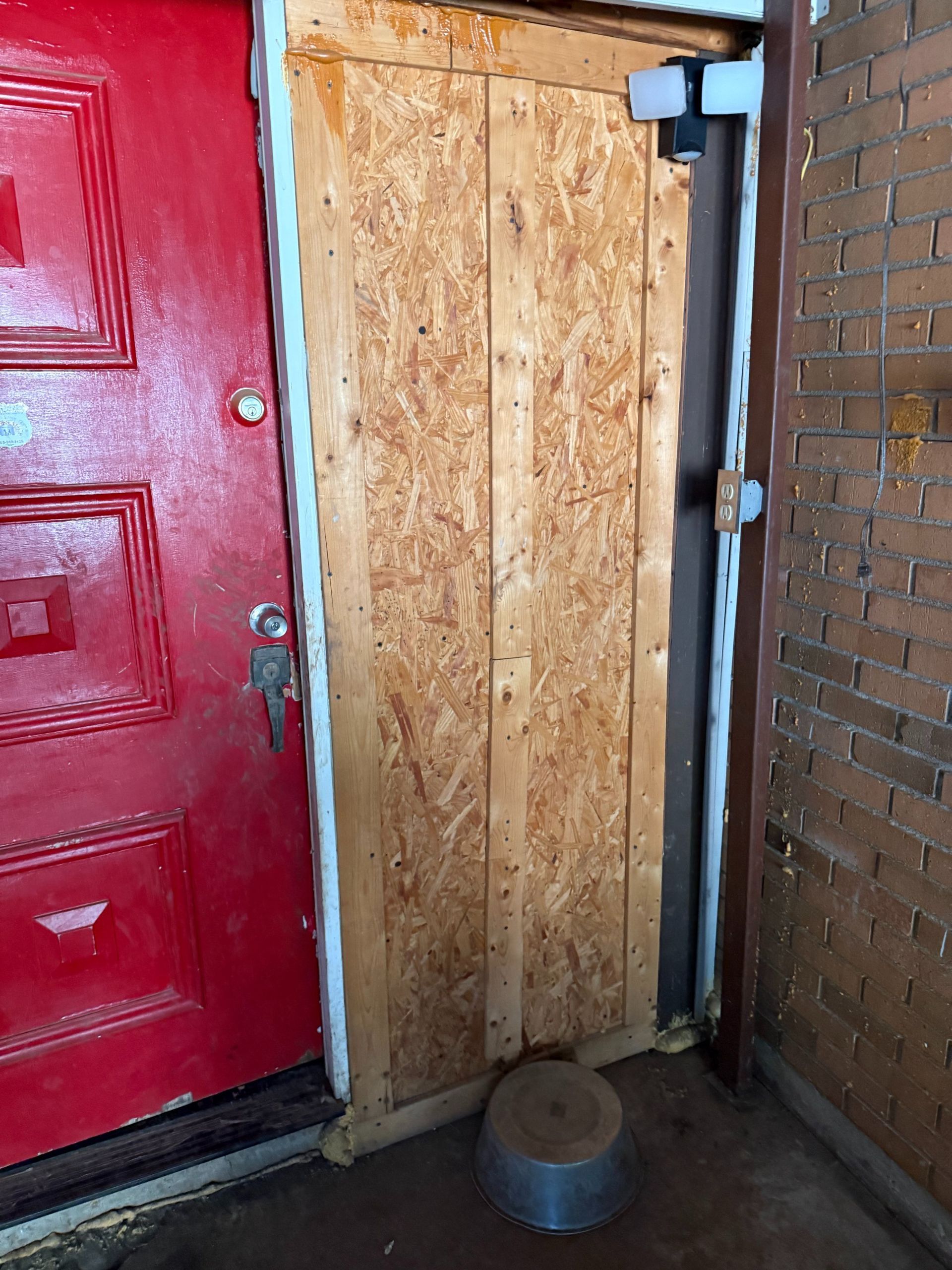 Red door next to a plywood-covered doorway. A metal hinge is visible.  A metal bucket sits on the floor.