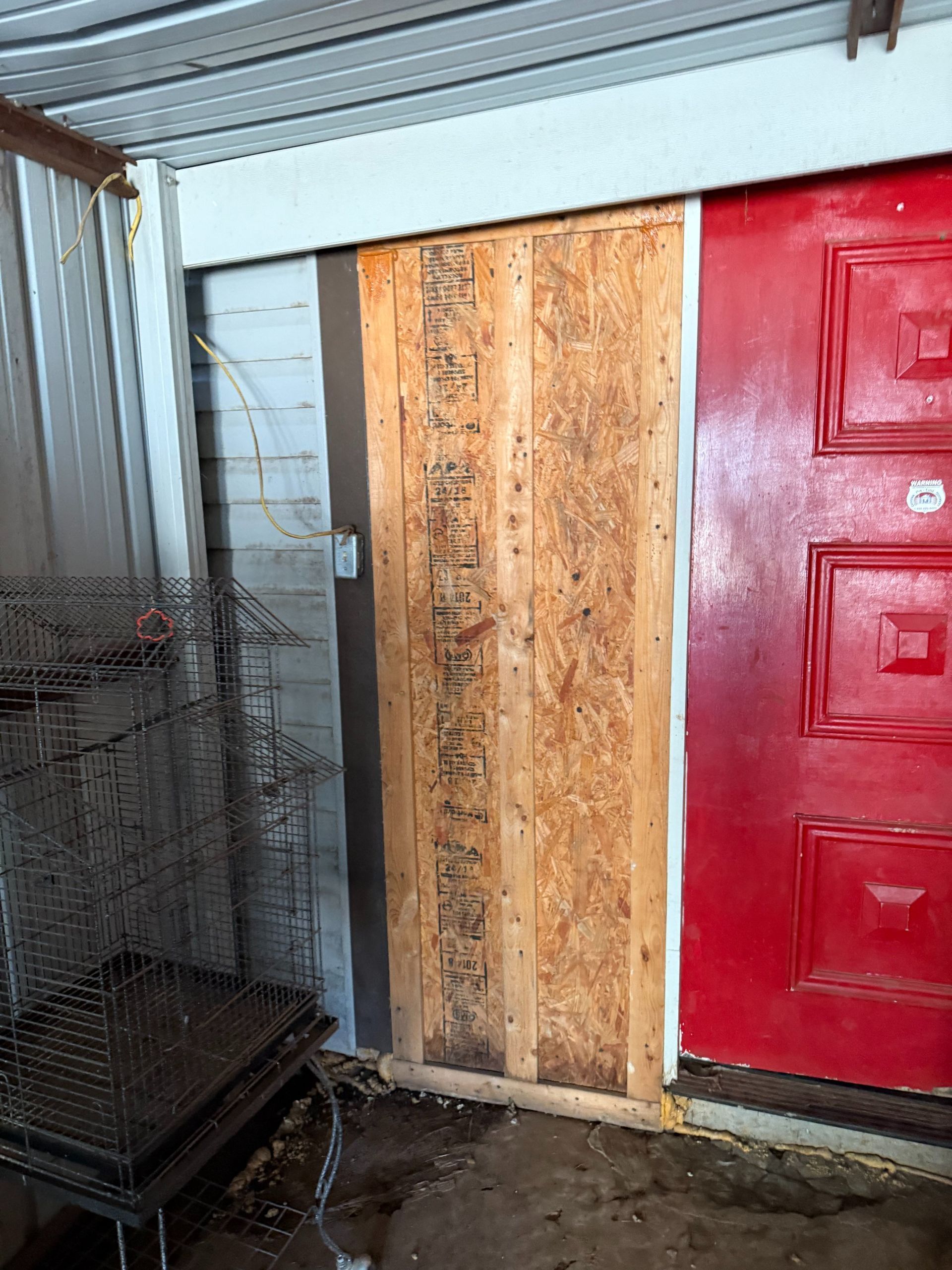 A wooden panel covers part of a doorway next to a red door.  OSB board, mounted on a white wall.