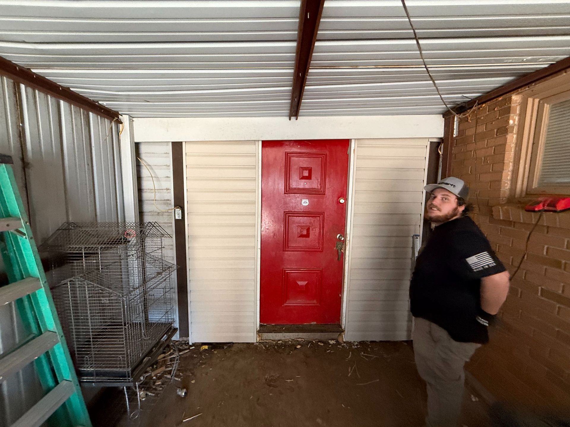 Man standing by a red door under a corrugated roof. Beige siding, green ladder, and metal cages are visible.