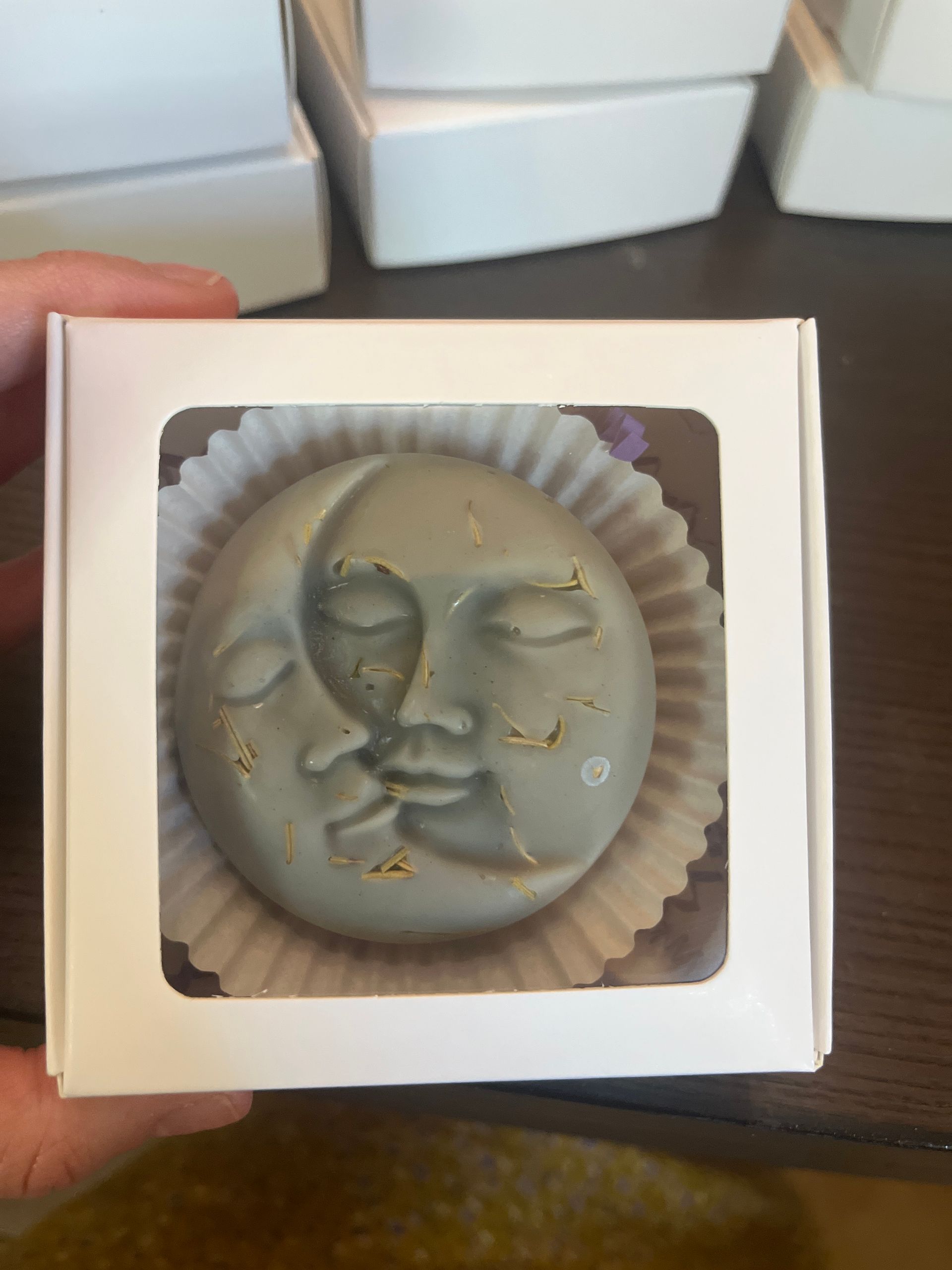 A person is holding a lotion bar in a box with a moon face on it.