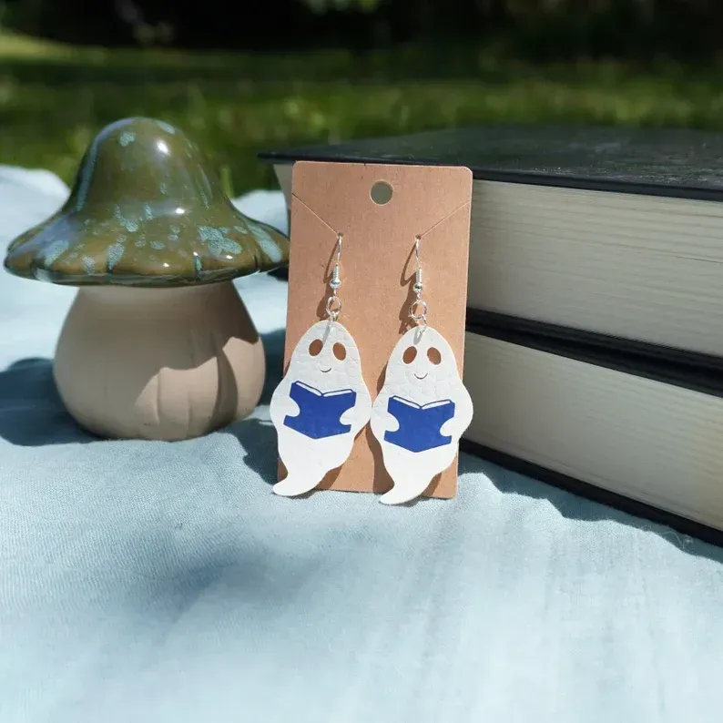 A pair of ghost earrings are on a card next to a mushroom and books