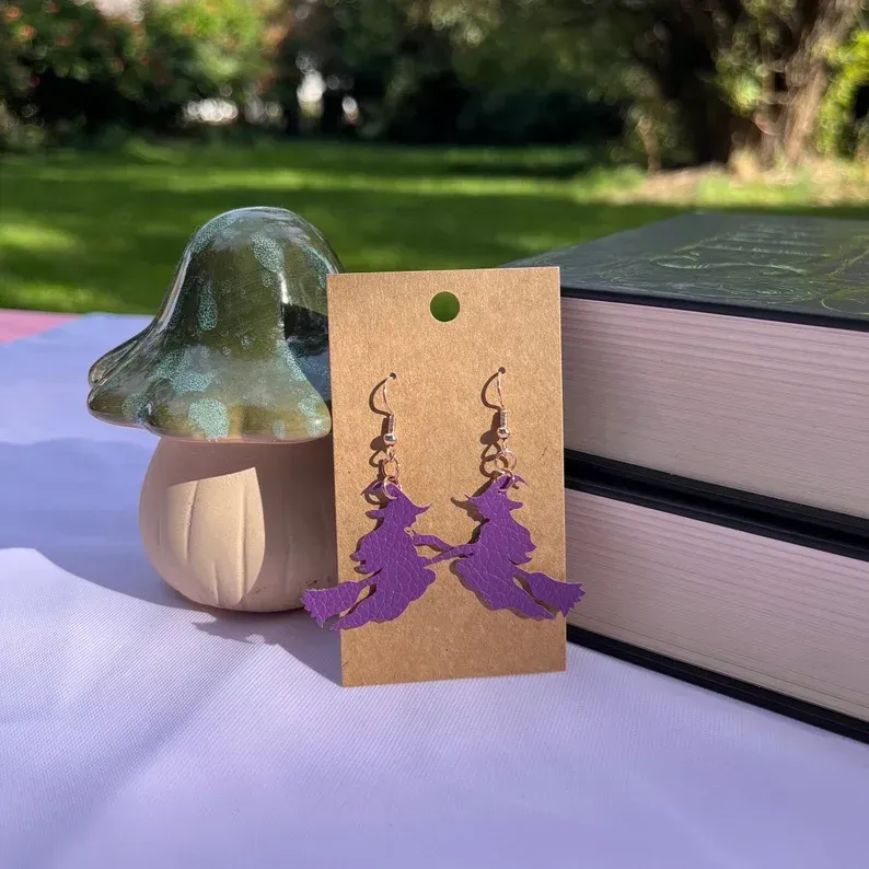 A pair of purple witch earrings are sitting on a table next to a mushroom and books.