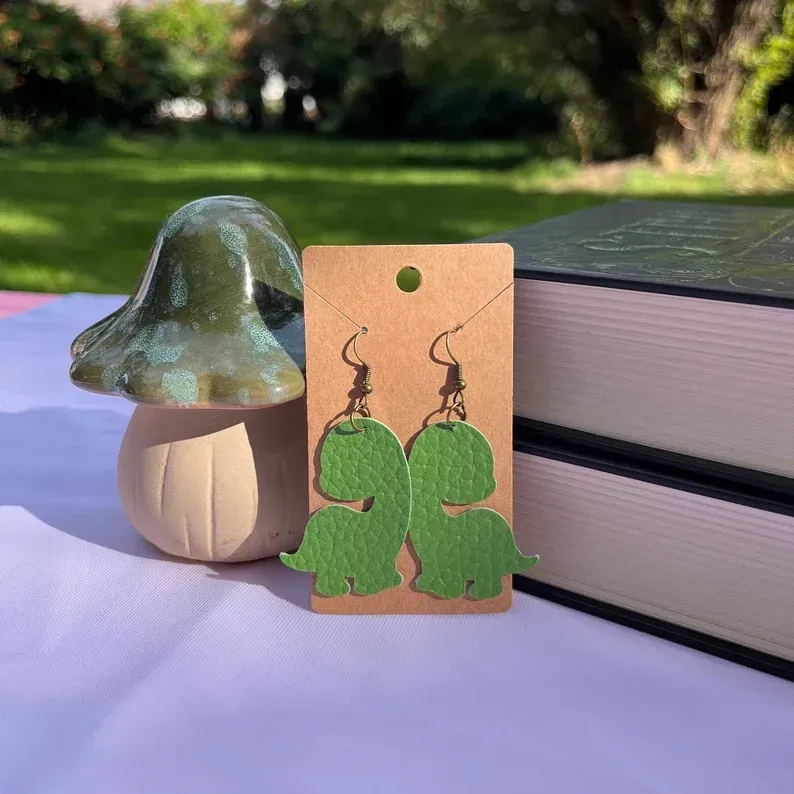 A pair of green dinosaur earrings are sitting on a table next to a mushroom.