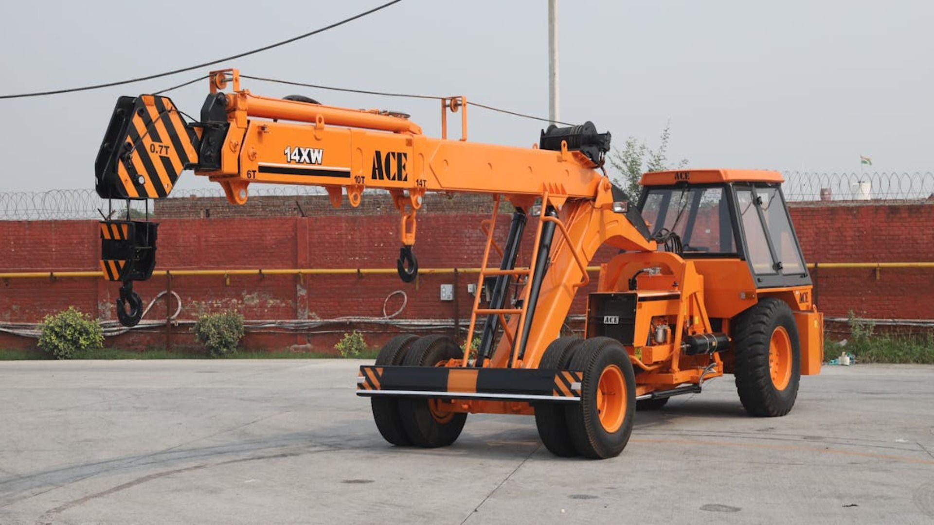 Grúa móvil ACE naranja sobre una superficie de hormigón. Brazo de grúa extendido, con gancho a rayas negras y amarillas.