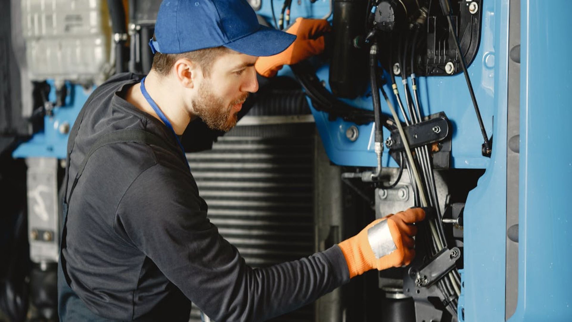Mecánico con gorra azul y guantes naranjas trabajando en maquinaria industrial.