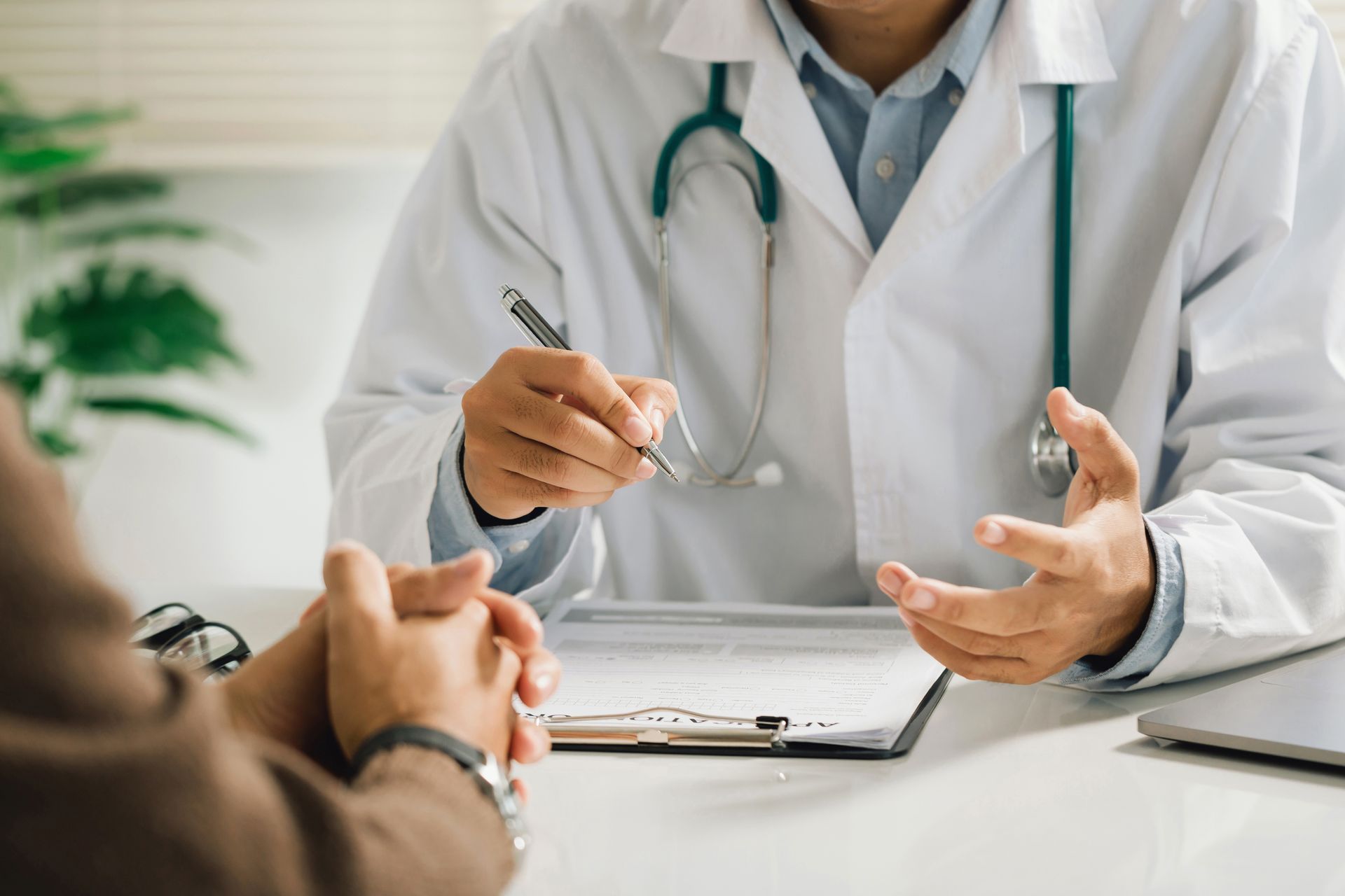 Ein Arzt unterhält sich mit einem Patienten in einer hellen Klinik und hält dabei einen Stift über ein Klemmbrett.