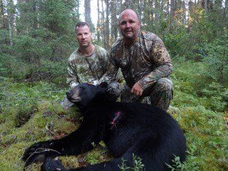 Two men are kneeling next to a black bear in the woods.