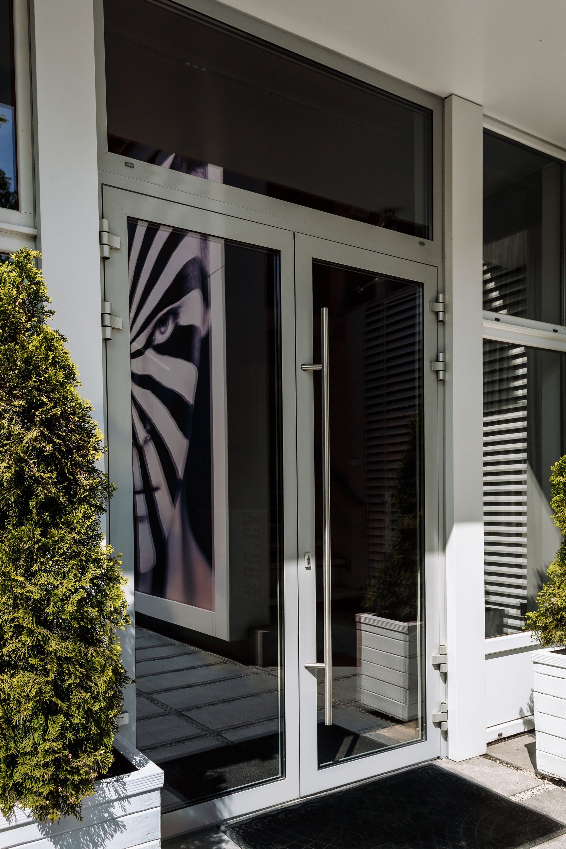 Una coppia di porte a vetri di colore chiaro con un disegno a zebra visibile attraverso il pannello di sinistra, fiancheggiate da piccoli alberi.