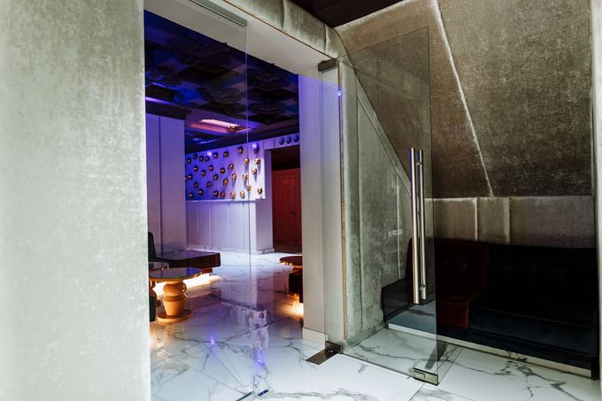 Uno spazio interno moderno e scarsamente illuminato, visibile attraverso una porta a vetri, caratterizzato da pavimenti in marmo, luci d'accento blu e posti a sedere.