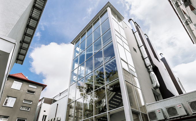 Un edificio moderno è caratterizzato da un'alta facciata continua in vetro che riflette il cielo, fiancheggiata da strutture bianche e condizionatori d'aria.