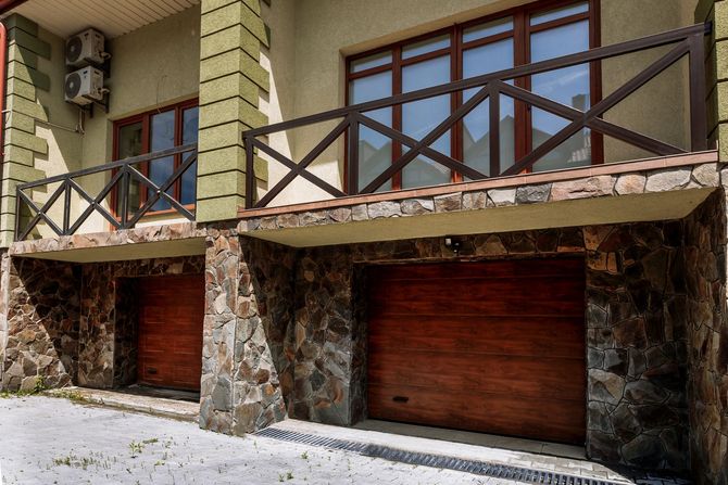 Due portoni da garage in legno scuro sotto un balcone con ringhiera marrone, in contrasto con una facciata in pietra e stucco chiaro.