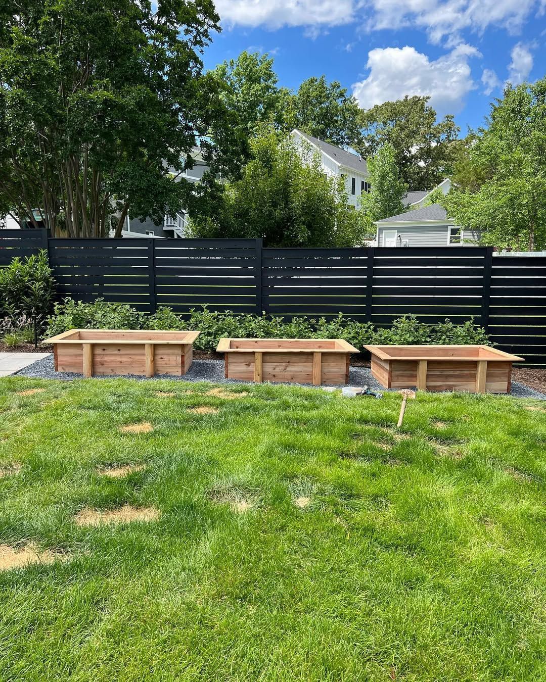 Three empty wooden raised garden beds in a grassy yard, with a dark horizontal fence and trees in the background.