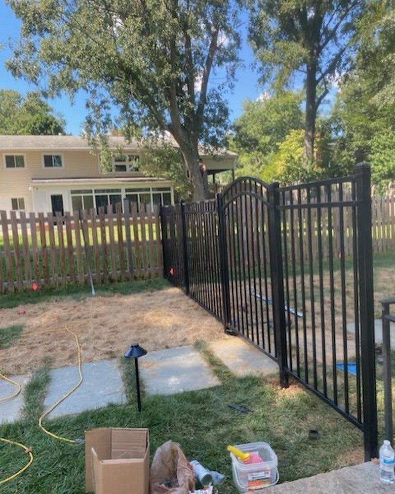 Black metal fence in a yard, leading to a gate. A house and trees are in the background.