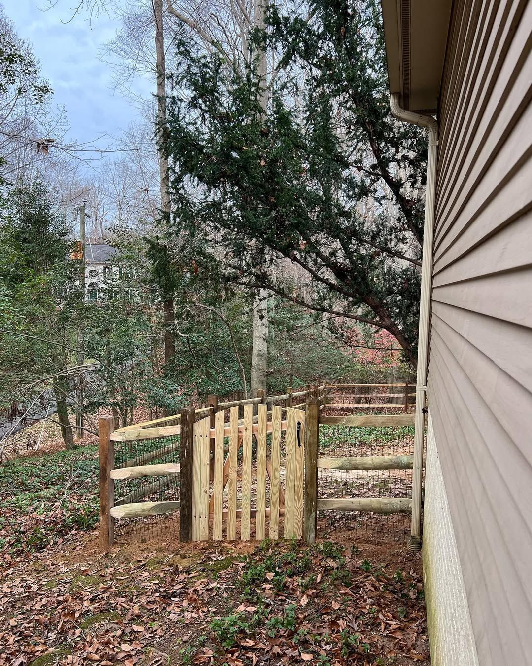 Wooden fence with gate next to a house; foliage and trees in the background.