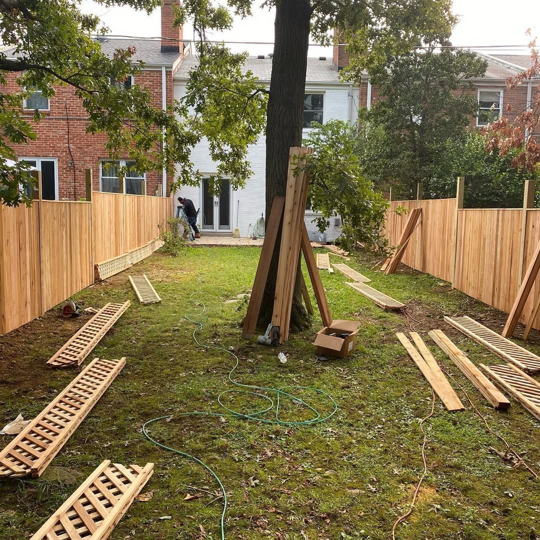 Backyard with a wooden fence under construction, scattered lumber, and a person working near a tree.