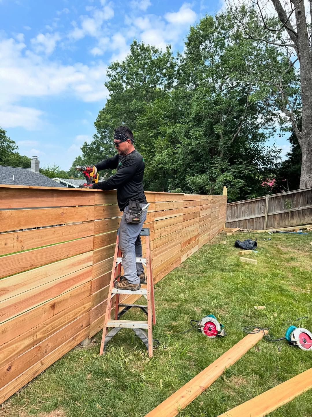 Man on ladder building a horizontal wood fence in a yard on a sunny day.