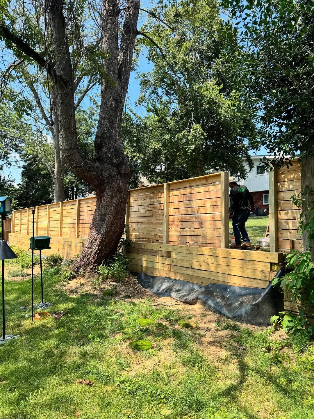 Wooden fence with a tree growing through it; person near open gate. Green grass and blue sky.