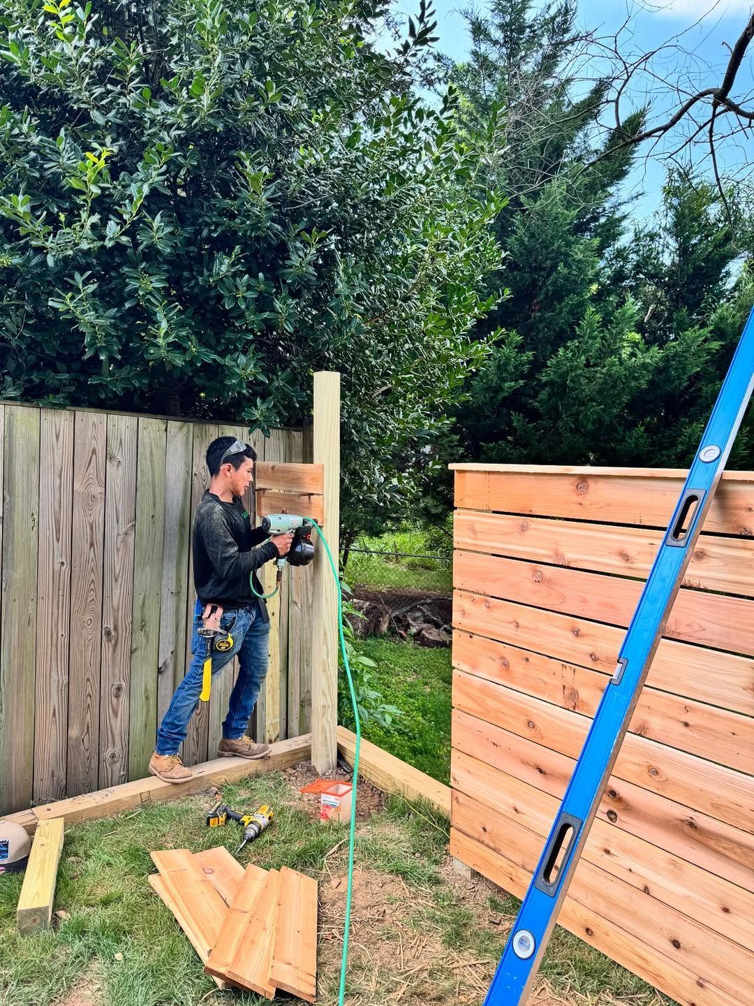 Man builds a wooden fence. He uses a nail gun on a post; lumber and tools are nearby, outdoors.
