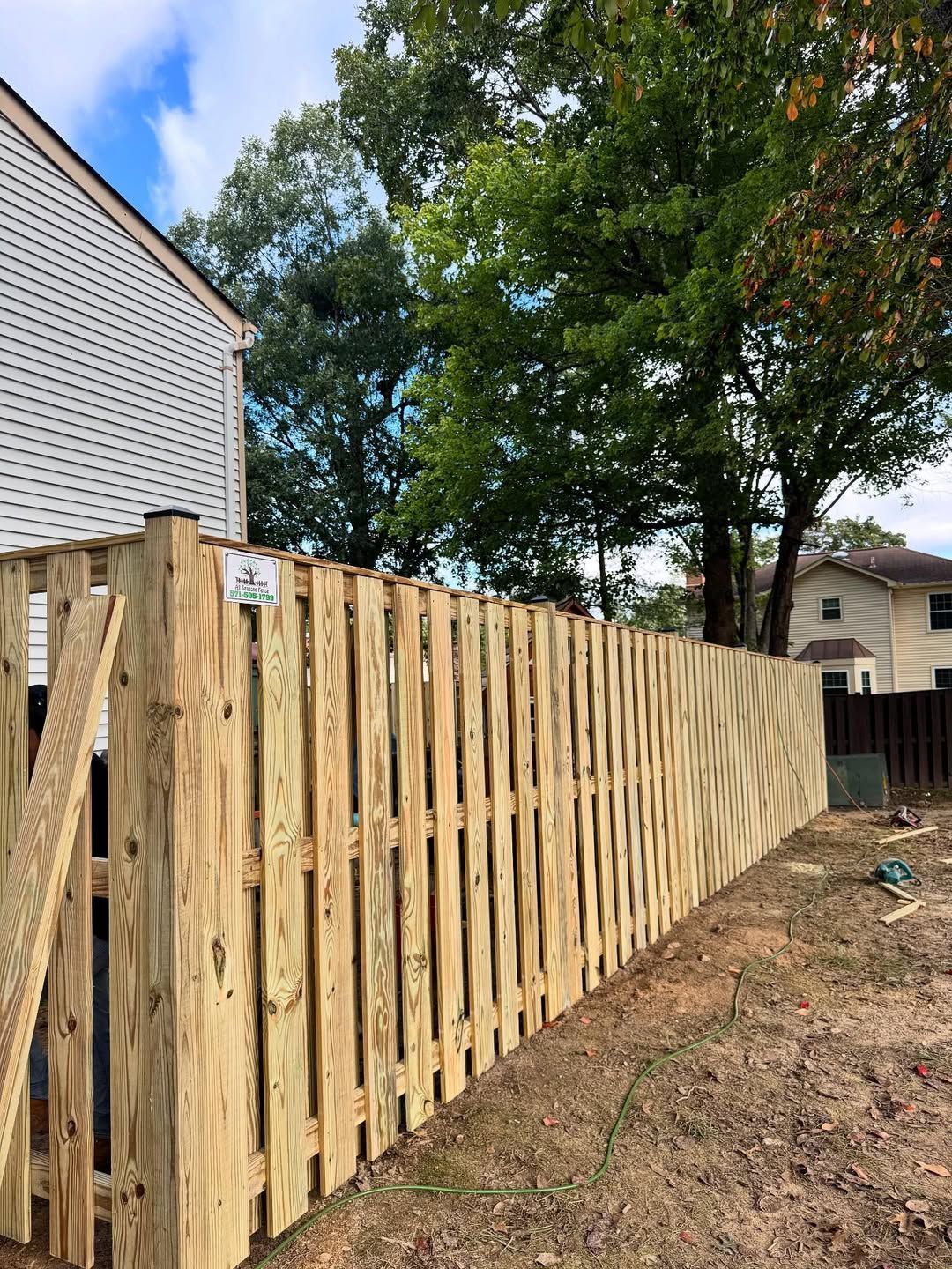 Newly built wooden fence in a yard, beside a white house and trees.
