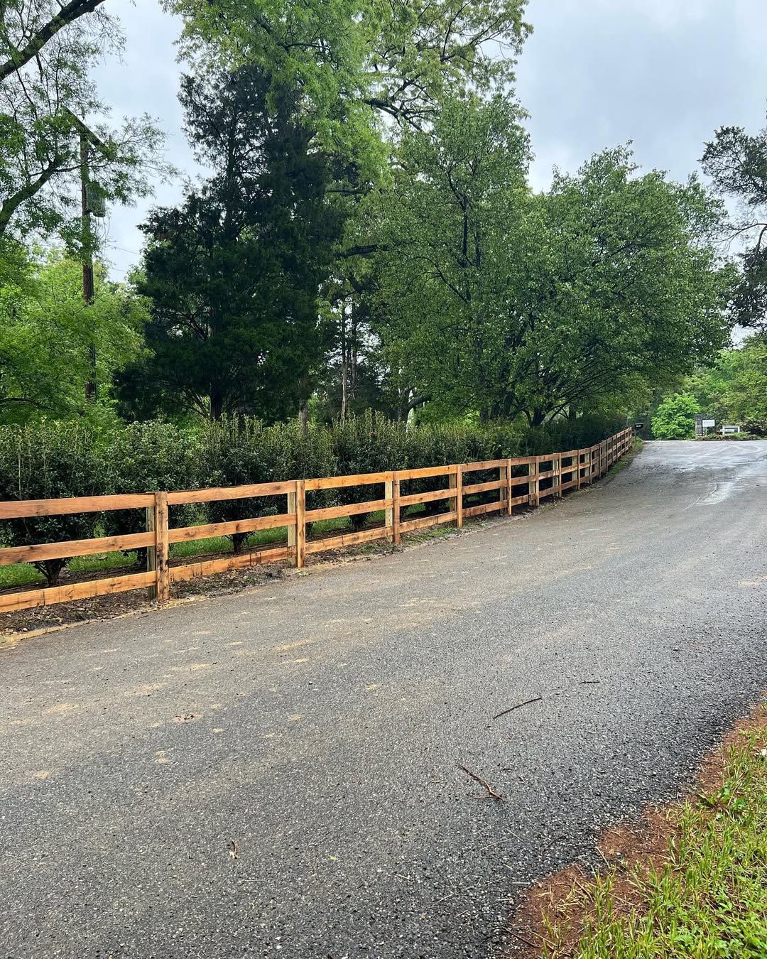 Gravel driveway curves past a wooden two-rail fence and green shrubbery, trees in the background.
