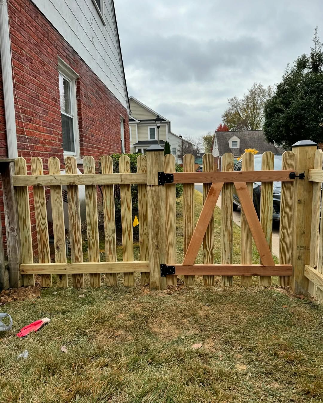 Wooden picket fence with a gate, next to a red brick building, cloudy day.