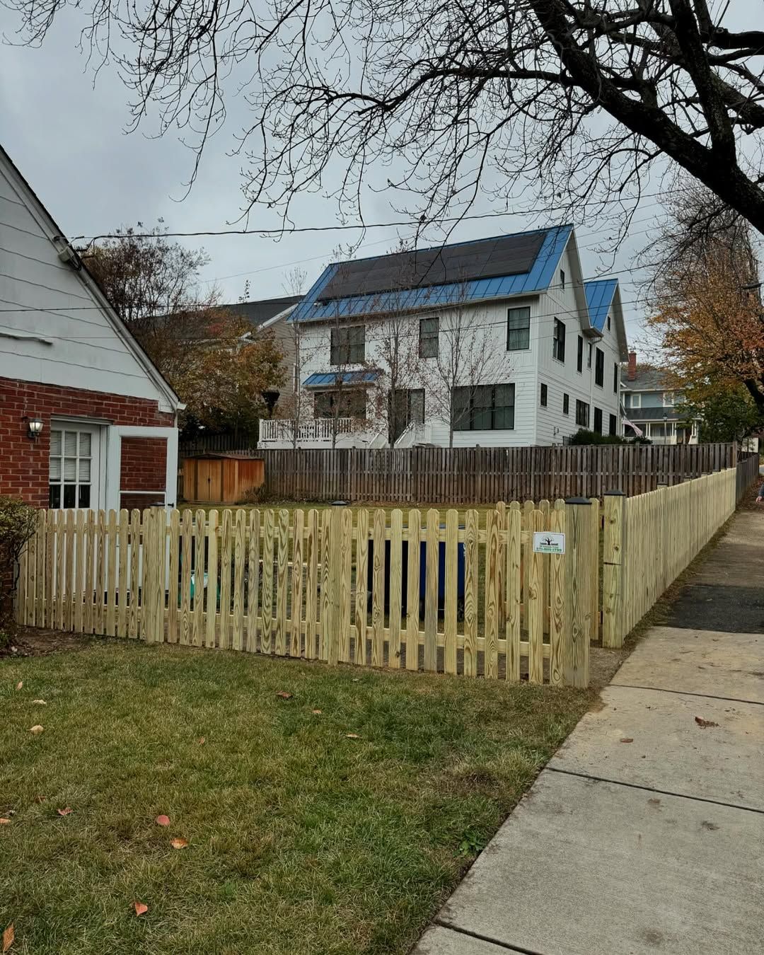 Wooden picket fence surrounds a grassy yard on a cloudy day. A house with solar panels is visible in the background.