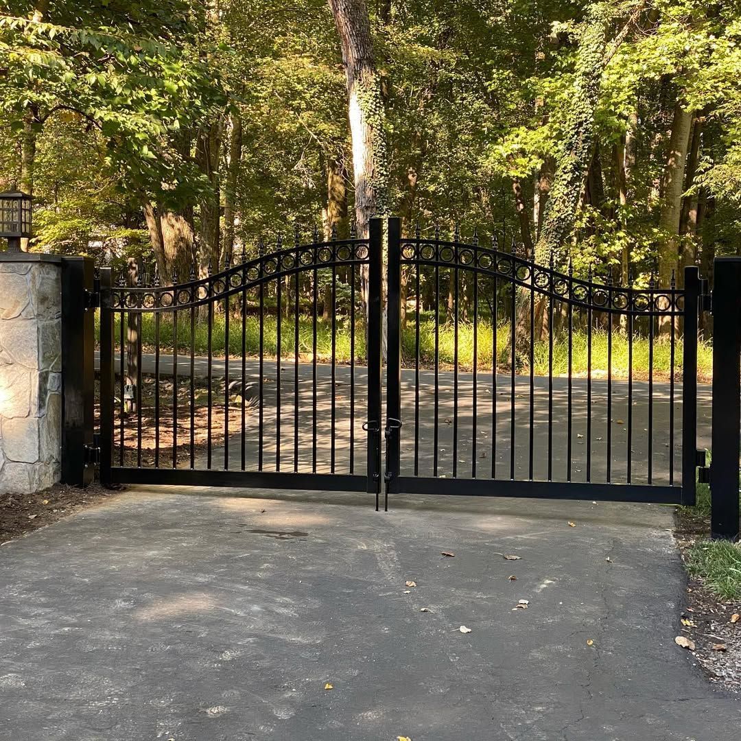 Black wrought-iron driveway gates with arched tops, set in a paved entrance with a wooded background.