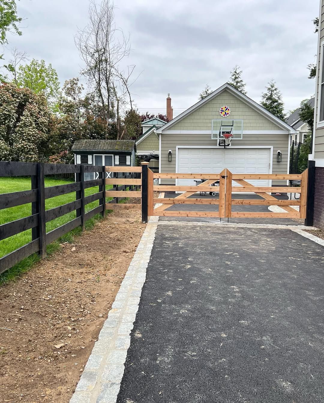Wooden gate entrance to a driveway, bordered by a black fence and a house with a basketball hoop.