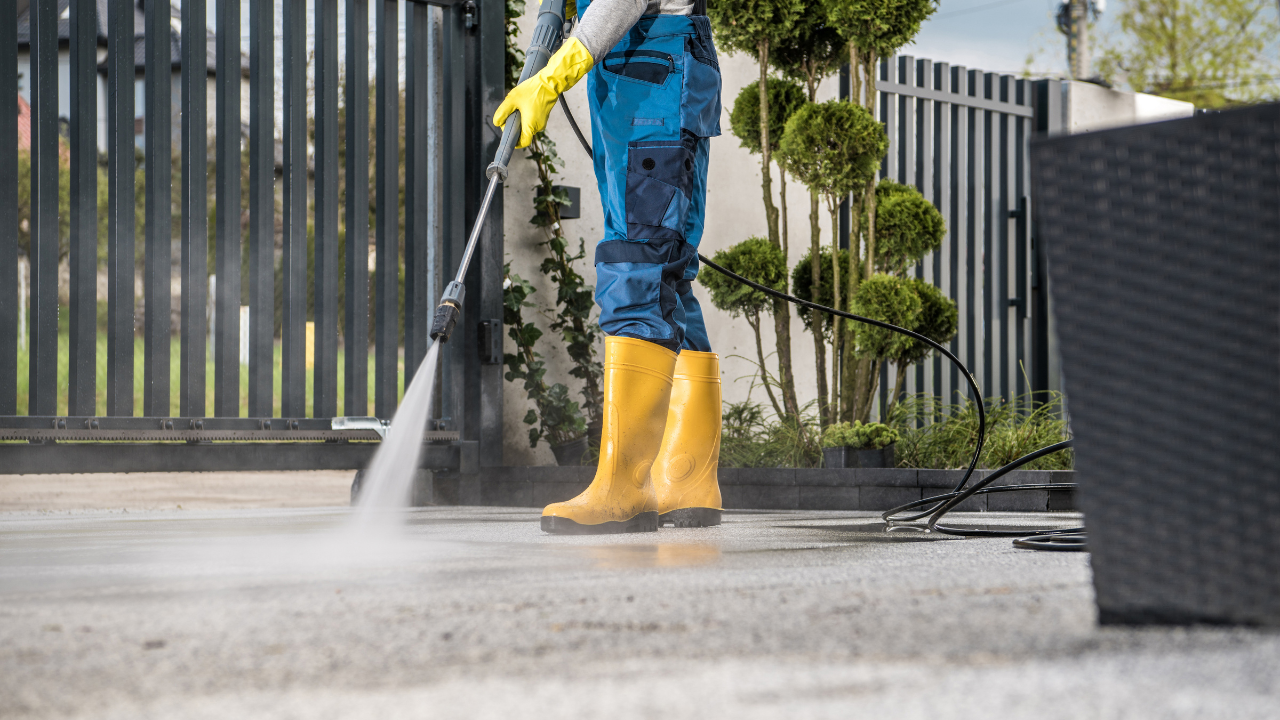 Person in yellow boots and gloves pressure washing a concrete patio near a fence.