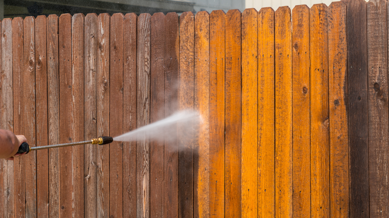 A person pressure washing a wooden fence, revealing clean, bright wood.