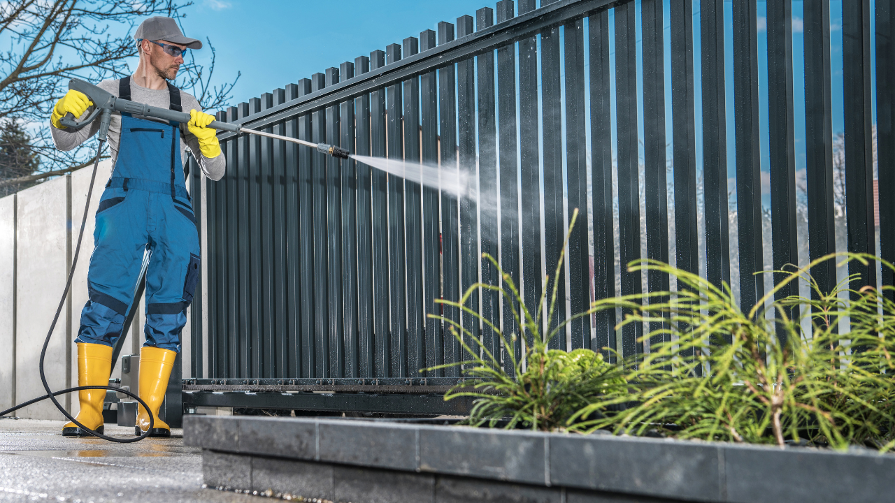 Man power washing a dark gray metal fence, wearing blue overalls and yellow boots.