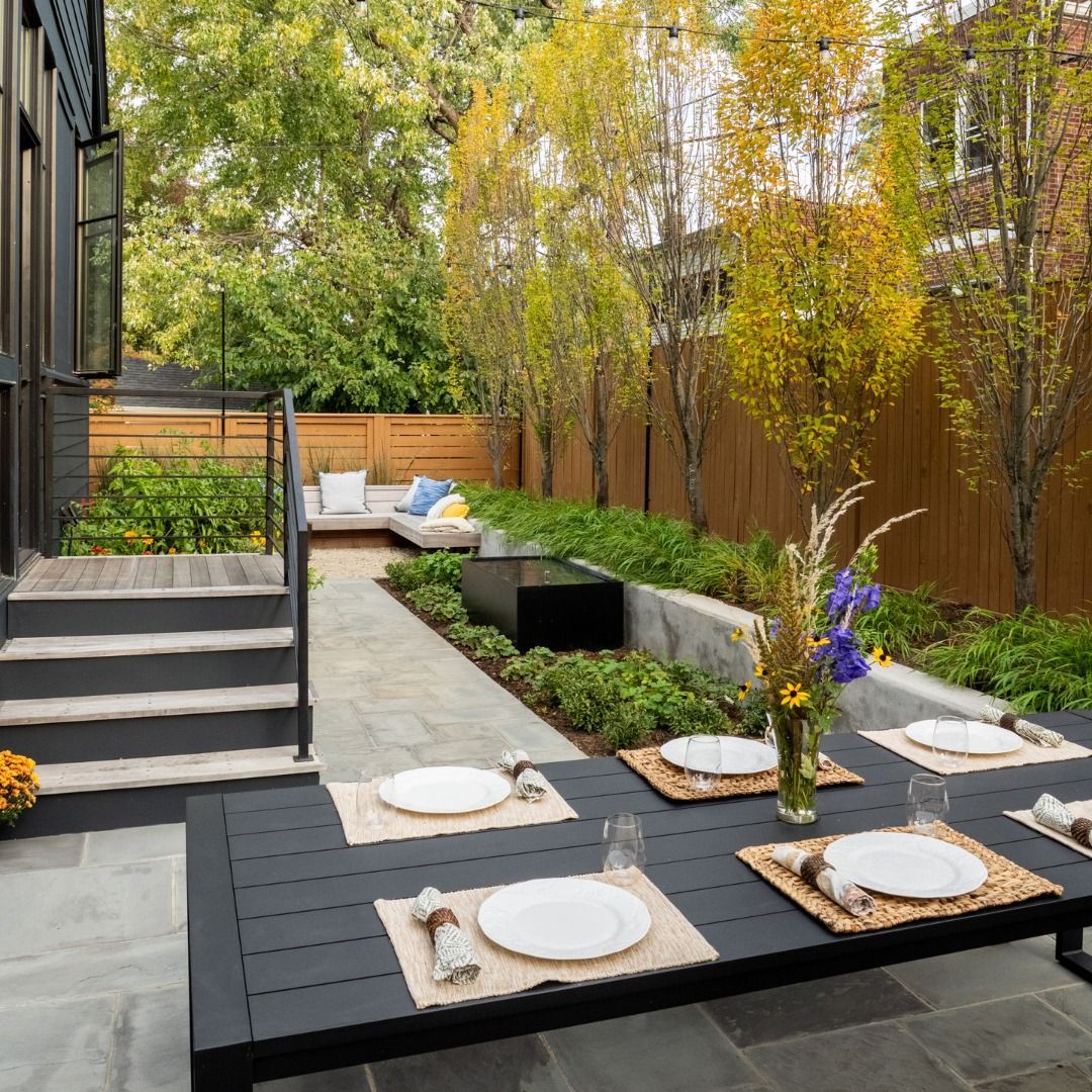 Outdoor dining space with dark table set for a meal, garden with trees and seating area in the background.