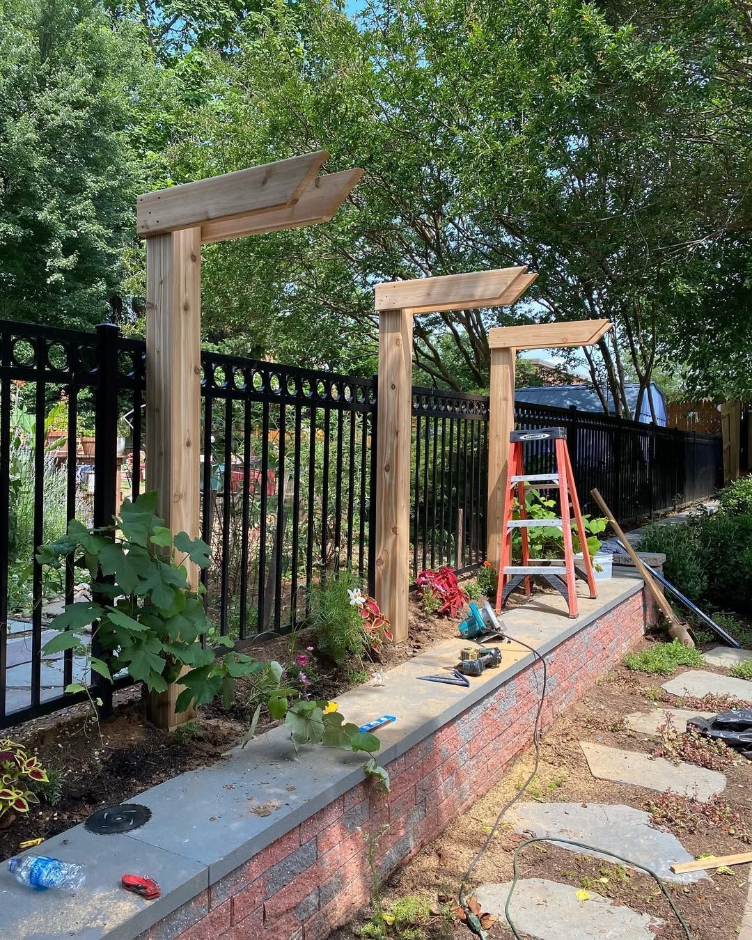 Wooden pergola posts being built along a black fence on a brick wall, with a ladder and tools.