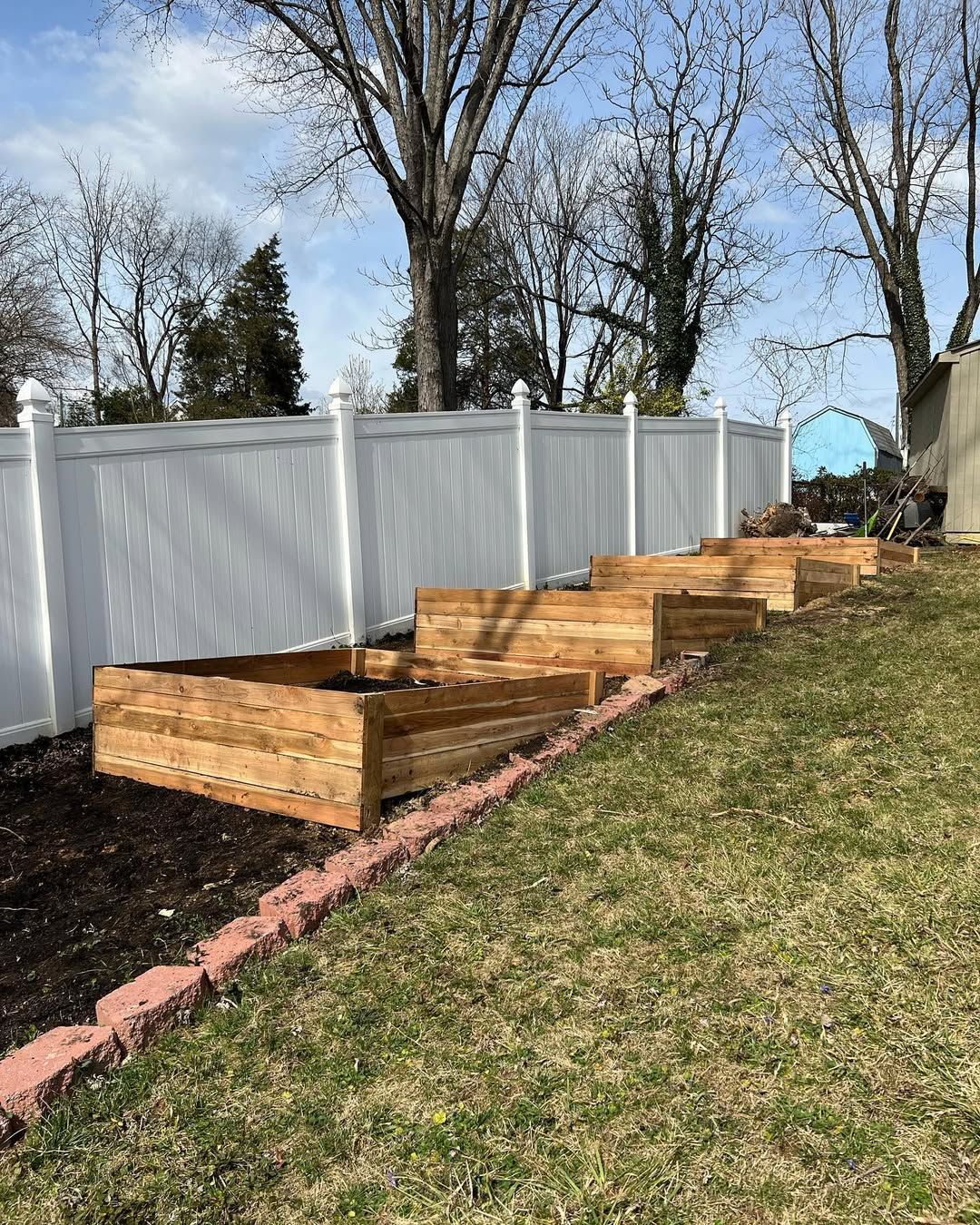 Wooden raised garden beds in a row on a grassy slope, with a white fence in the background.
