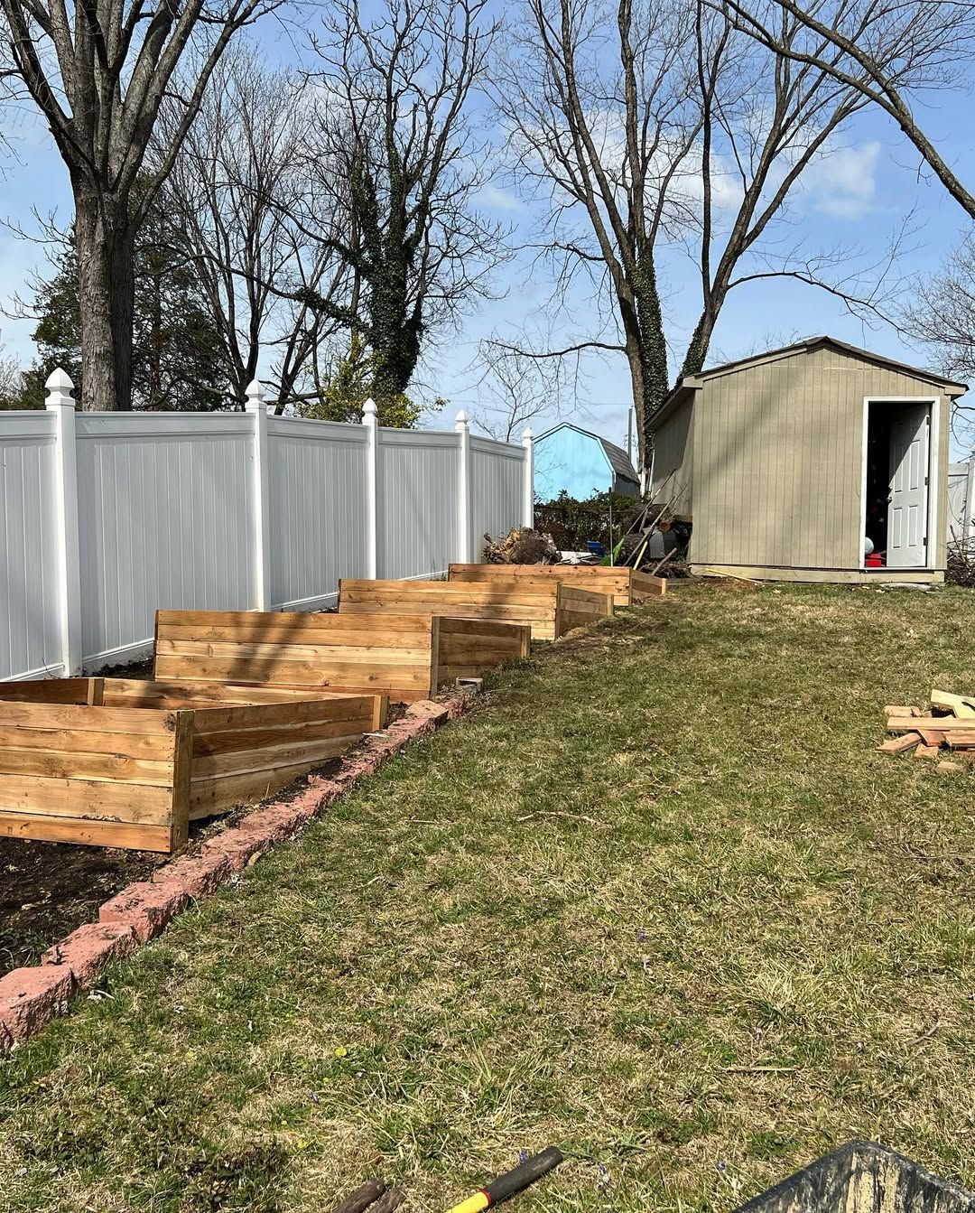 Wooden terraced garden beds on a sloped lawn, beside a white fence and a shed.
