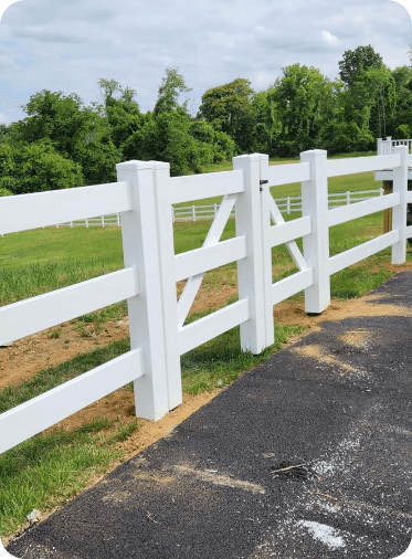 White fence with a gate in front of a green field and trees.