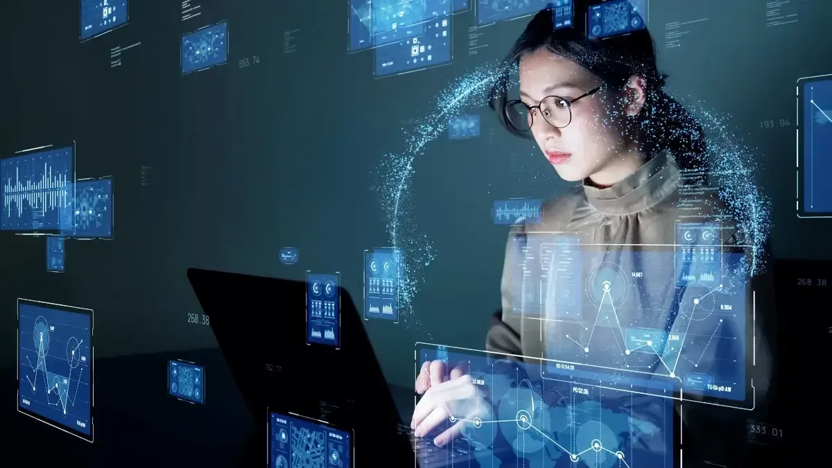 Woman in glasses working on a computer surrounded by floating digital data displays. Blue hues dominate.