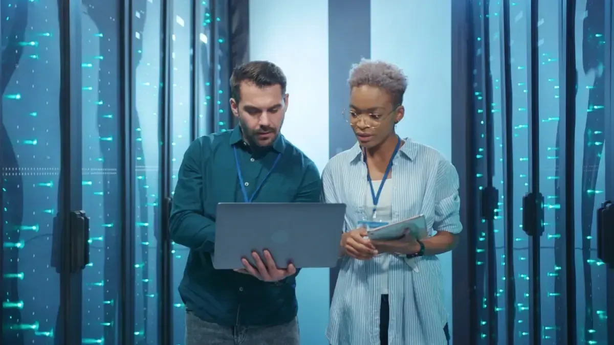 Two technicians using a laptop and holding paperwork in a server room with blue lights.