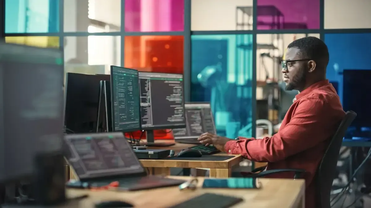 Man coding at multiple computer screens in office with colorful windows.