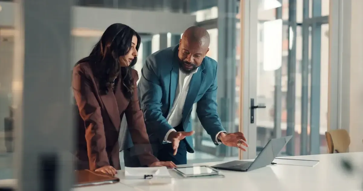 Two colleagues in business attire consult a laptop on a desk in a brightly lit, modern office.