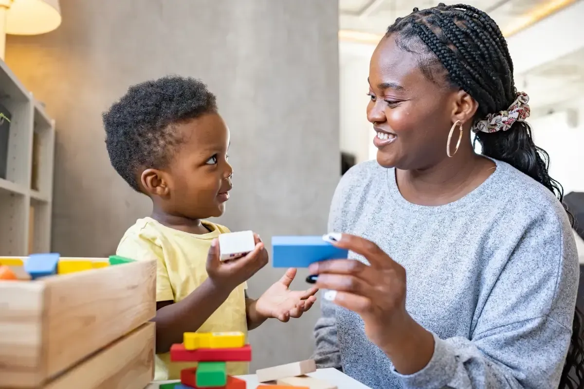Woman smiles while playing with a child, building with wooden blocks. Bright indoor setting.