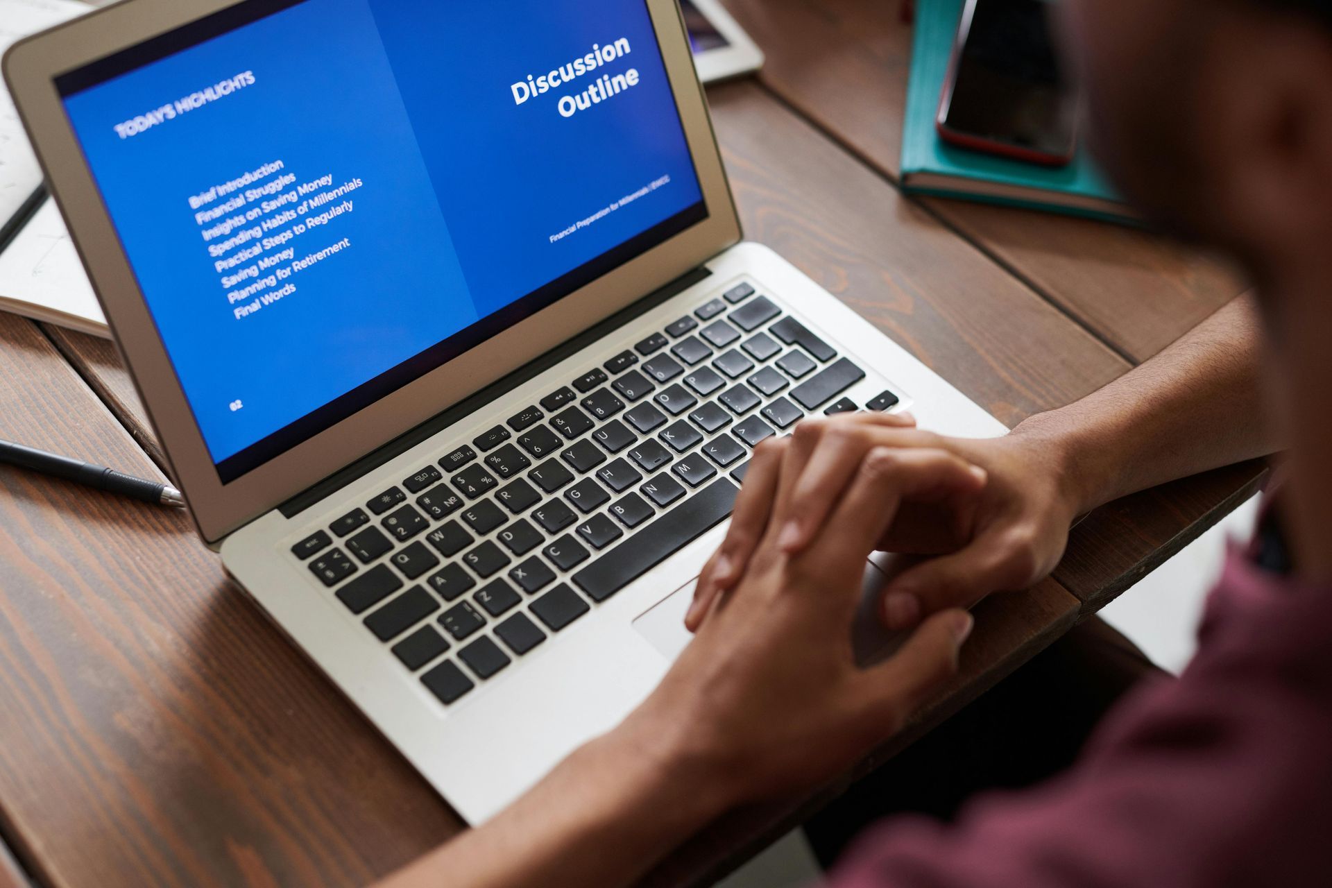 Person using a laptop, hands clasped, at a wooden table. The screen displays a blue presentation.