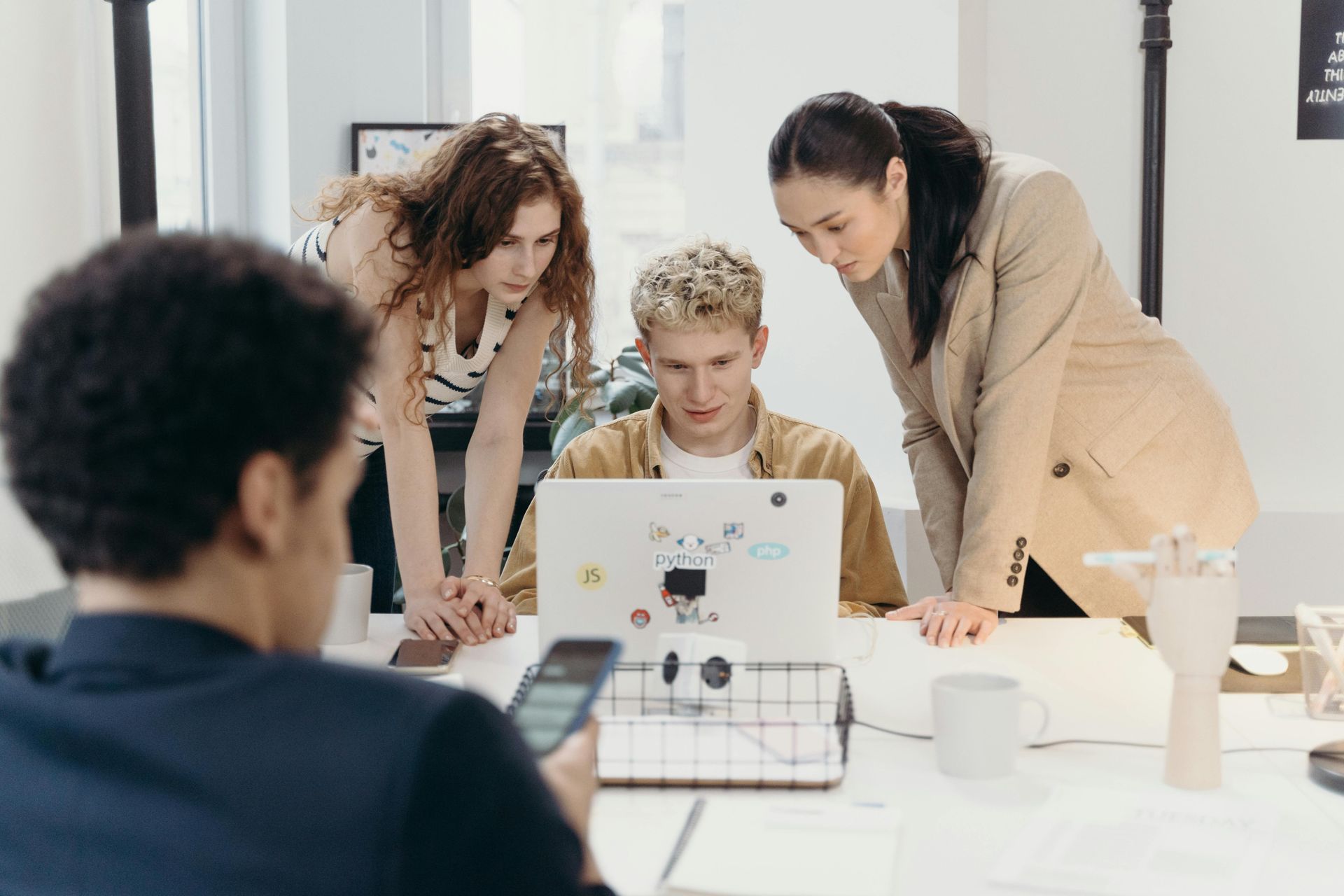 Four diverse colleagues collaborate at a desk, looking at a laptop and phone. Office setting, focused.