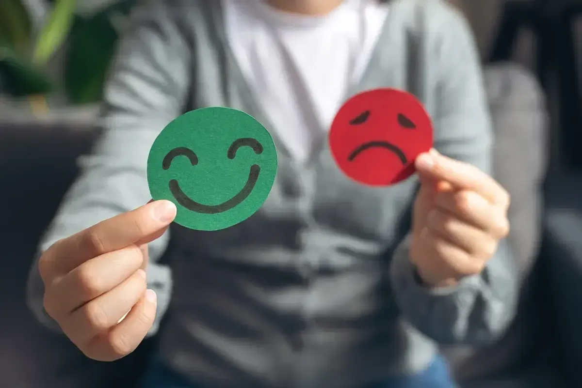 Person holding a green smiling face and a red frowning face, symbolizing positive and negative emotions.