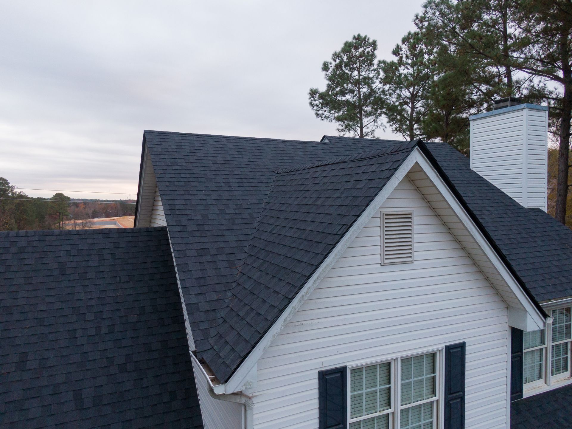 Dark blue shingle roof on a white house, chimney, and trees in the background under a cloudy sky.