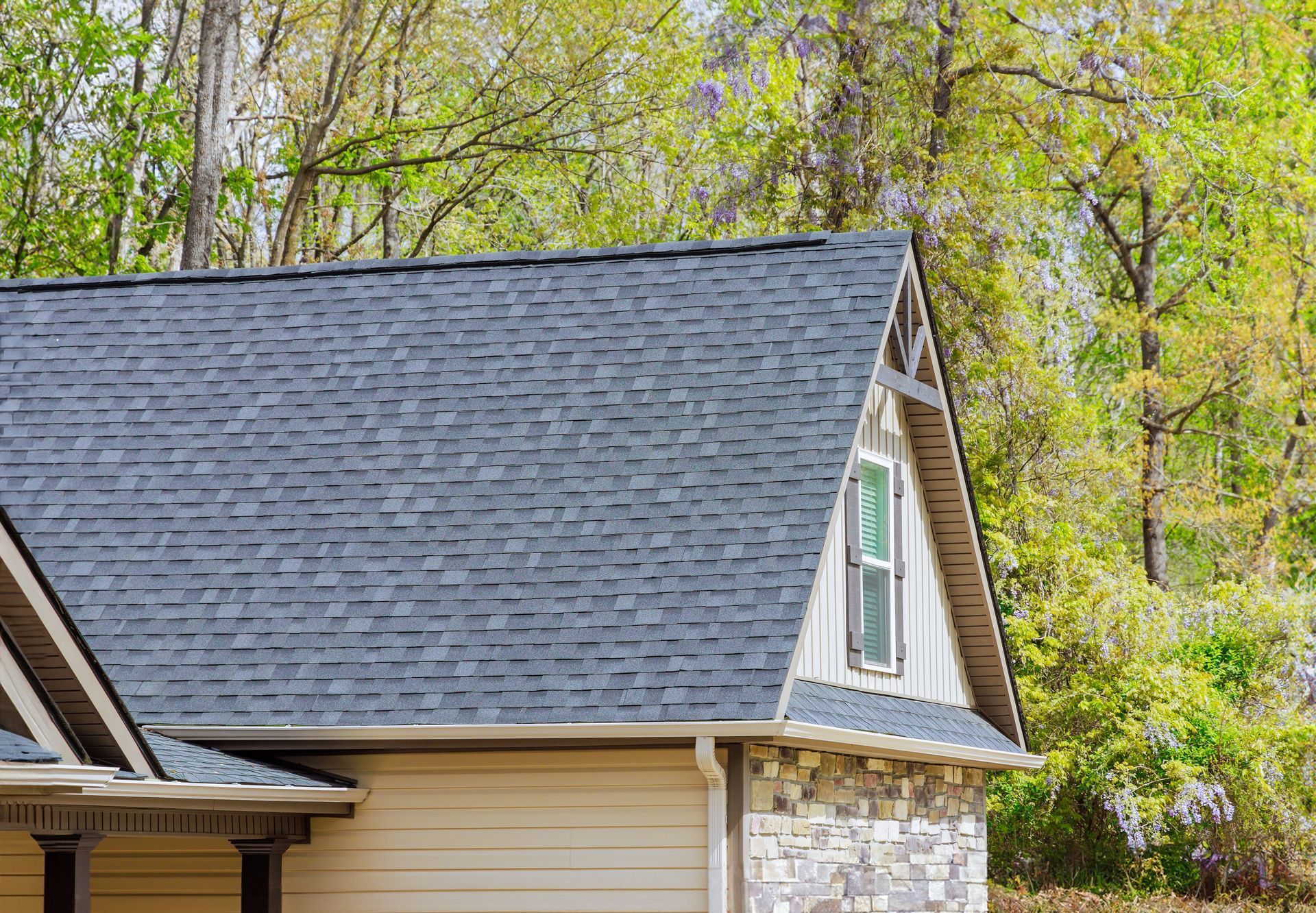 House with gray shingled roof, beige siding, stone facade, and trees in the background.