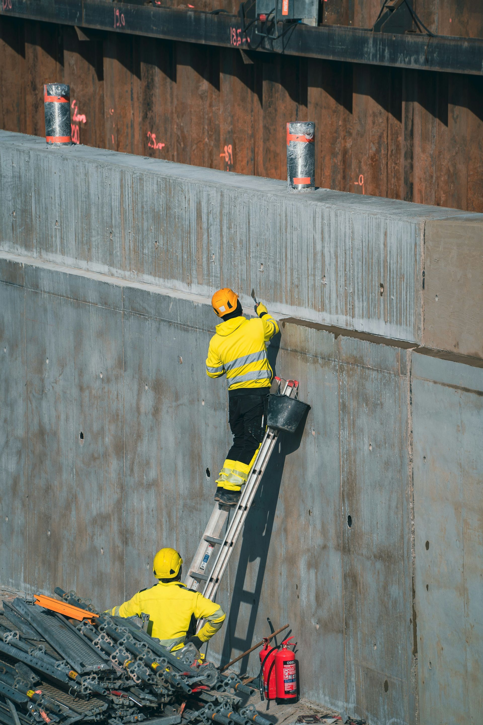 Two construction workers in yellow vests repair a concrete wall, one on a ladder, the other below.