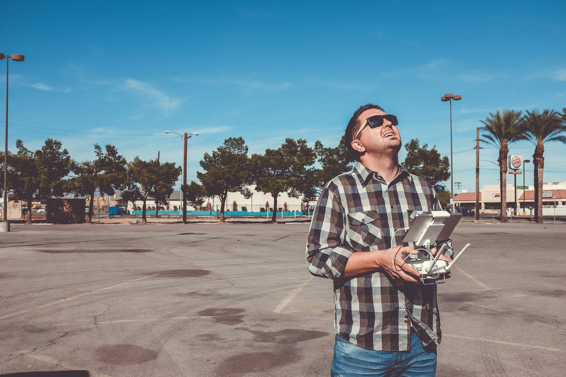 Man flying a drone in a parking lot, looking up at it. Wearing sunglasses, plaid shirt and jeans. Blue sky.
