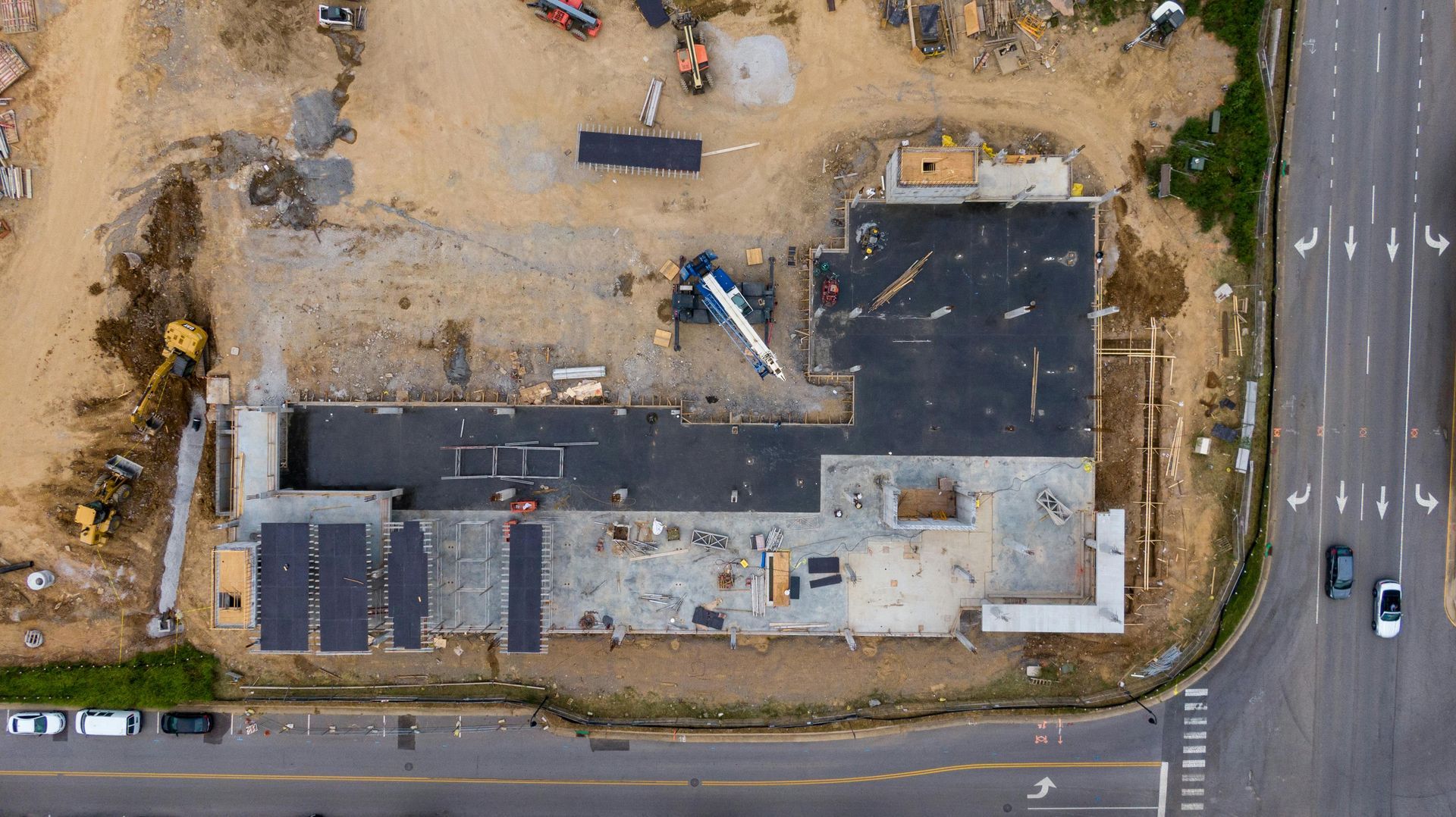 Aerial view of a construction site with foundation, crane, and materials. Next to a road with a few cars.