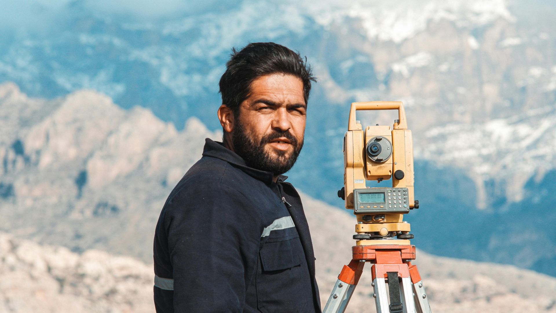 Man with beard using surveying equipment in a mountainous landscape.